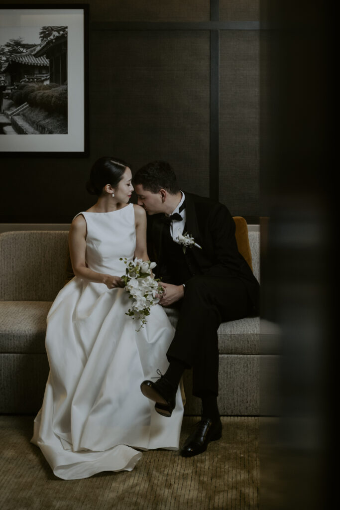 A bride and groom sitting on a couch in their Seoul hotel room.