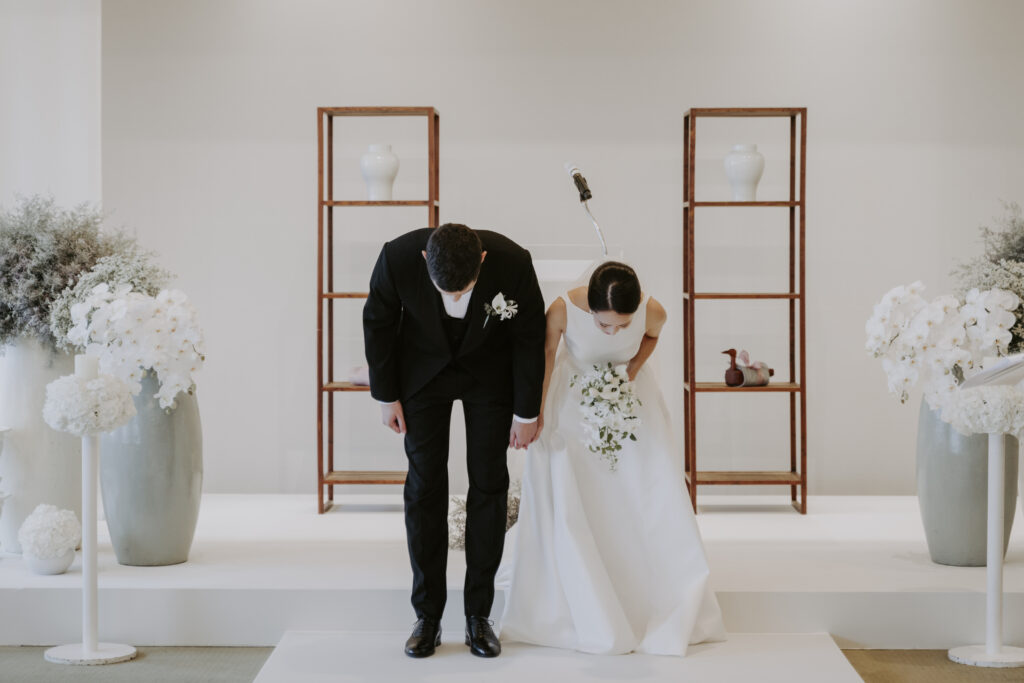 A bride and groom standing next to each other bow to their guests.