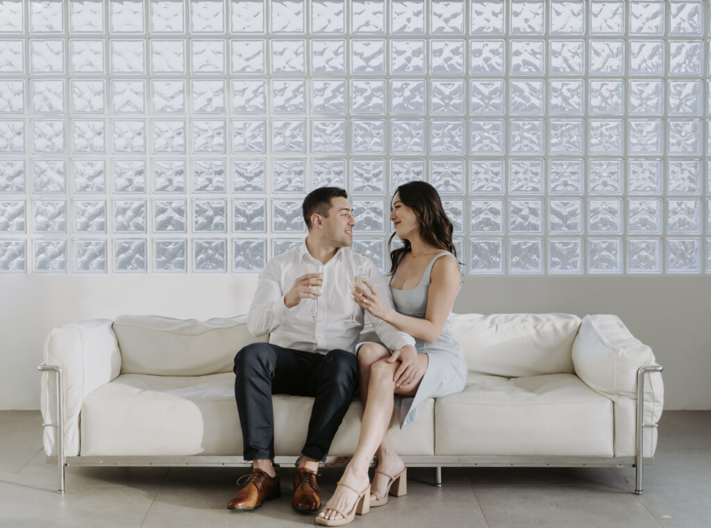 An engaged couple sitting on a couch in front of a wall.