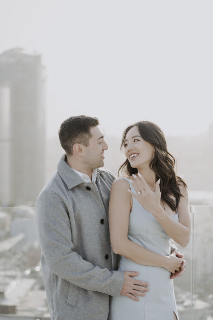 An engaged couple standing on a rooftop overlooking the city.