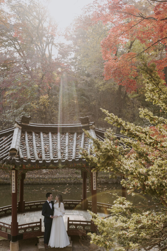 A bride and groom eloping in Seoul, posing in front of a pagoda adorned with autumn foliage.