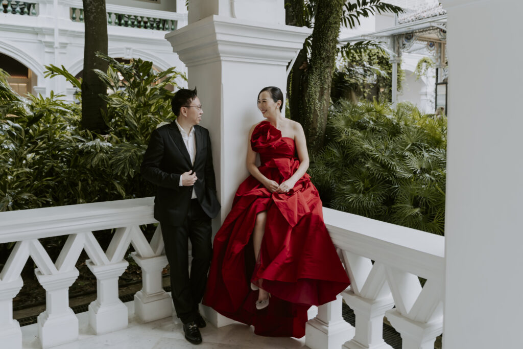 A bride and groom in a red dress standing on a balcony at Raffles Hotel Singapore.
