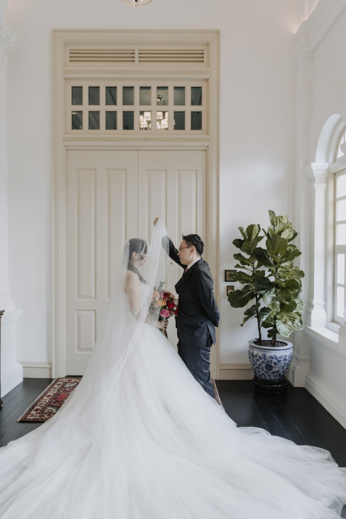 A bride and groom pose elegantly in front of the lavish grand ballroom at Raffles Hotel Singapore on their wedding day.