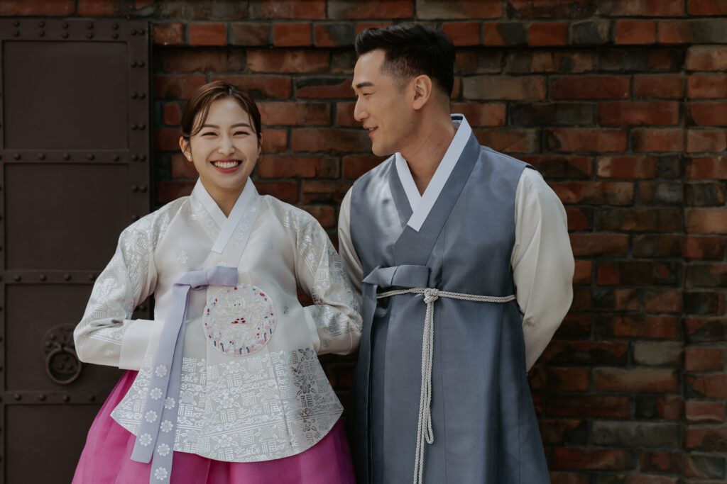 A couple in traditional korean clothing standing next to a brick wall in a hanok house. 