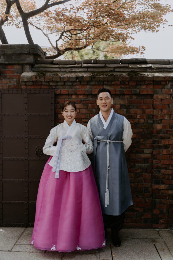 A couple in traditional korean hanbok standing in front of a brick wall of the hanok house. 