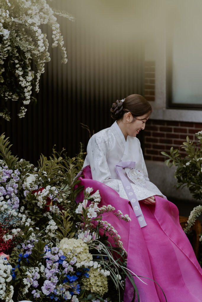 A bride in a pink hanbok sits on a flower bed.