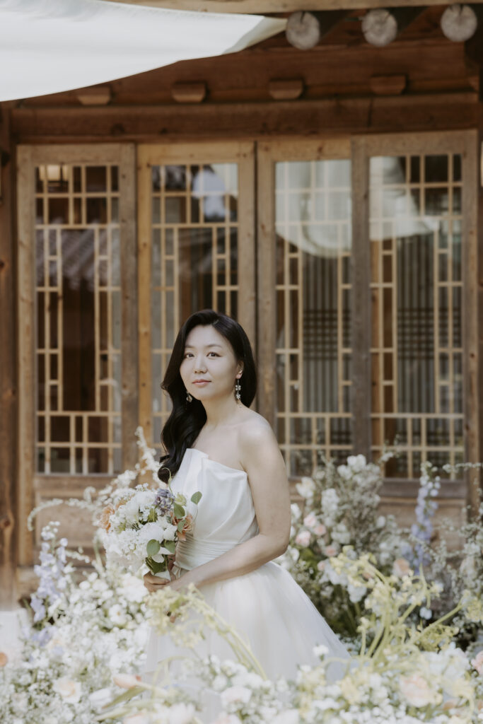 A bride in a white wedding dress posing in front of a hanok house.