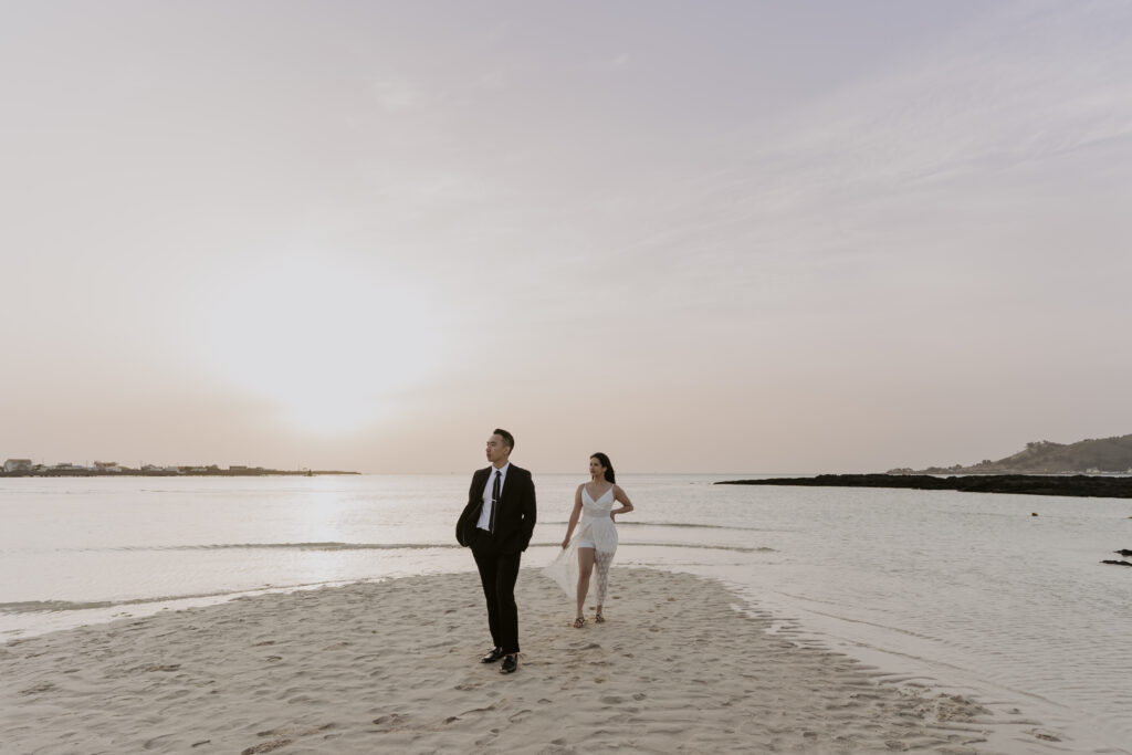 A bride and groom walking on the beach at sunset.