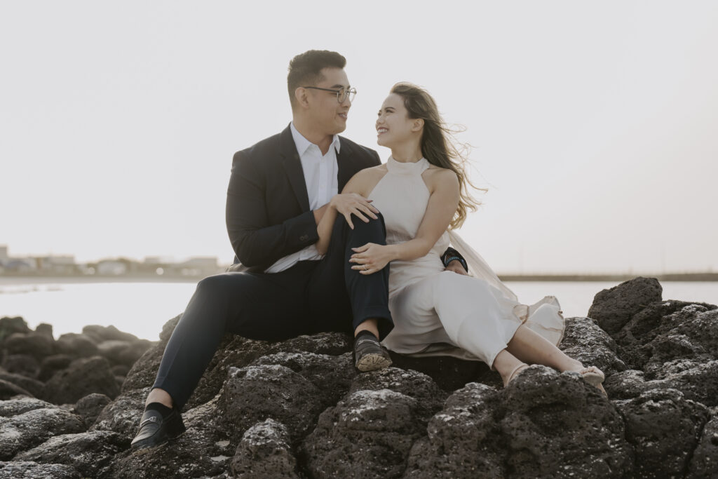 a bride and groom pose on the rocks at sunset during their beach pre-wedding photoshoot