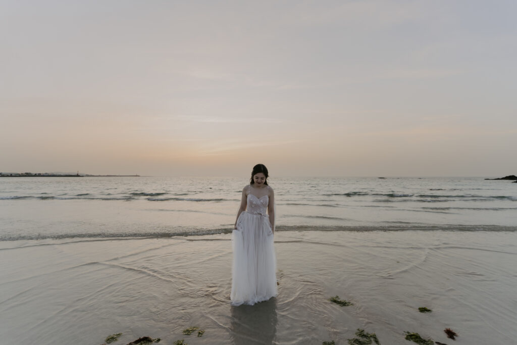 A woman in a white dress on Jeju Island, capturing moments during a pre-wedding photoshoot at sunset.