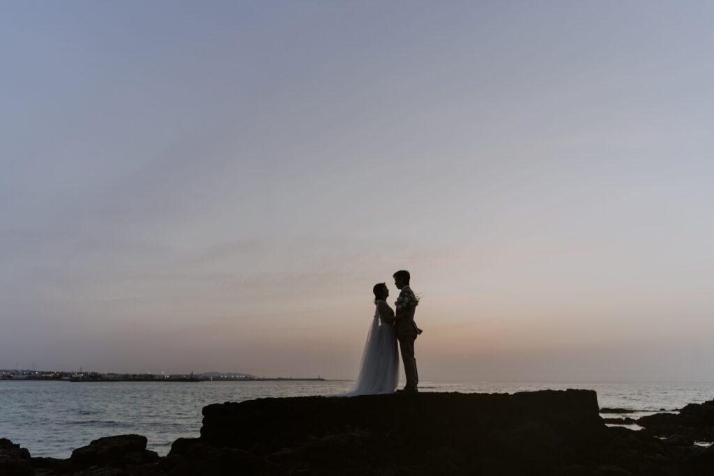 A pre-wedding photoshoot on Jeju Island captures a bride and groom standing on rocks at sunset.