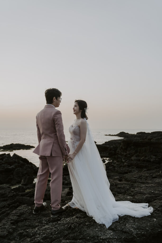 A pre-wedding photoshoot with the bride and groom standing on rocks at sunset on Jeju Island.