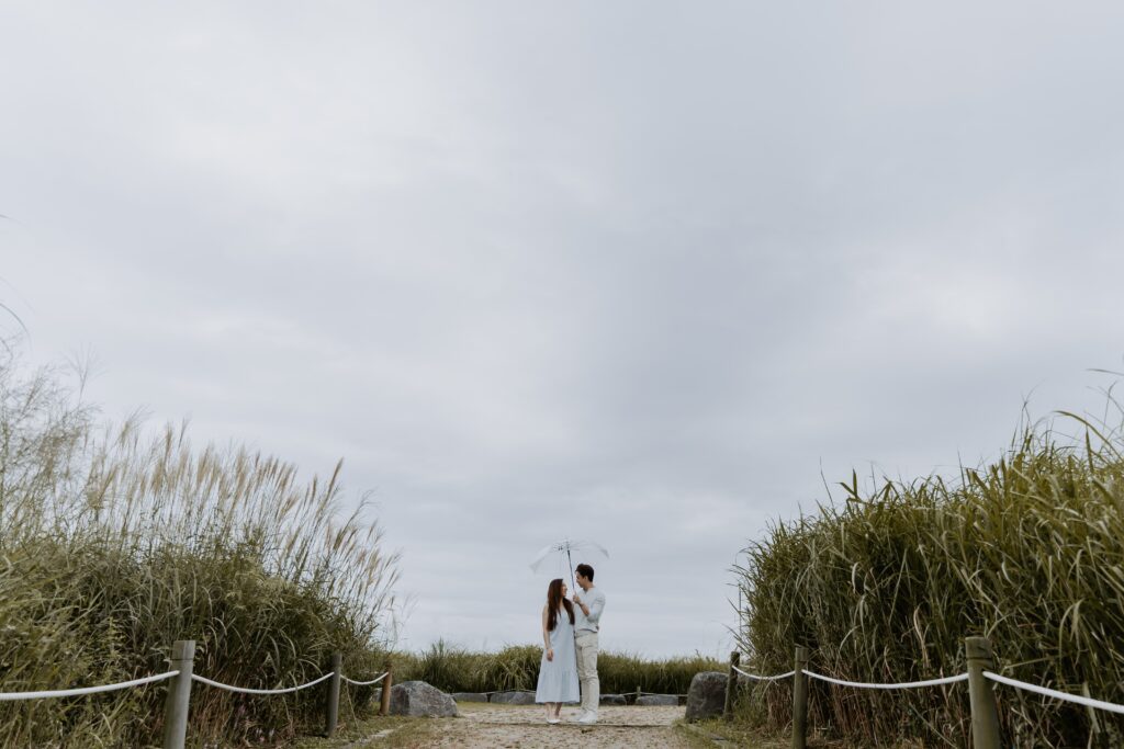 A bride and groom standing on a path with reeds in the background while it rain in Haneul Park, Seoul South Korea