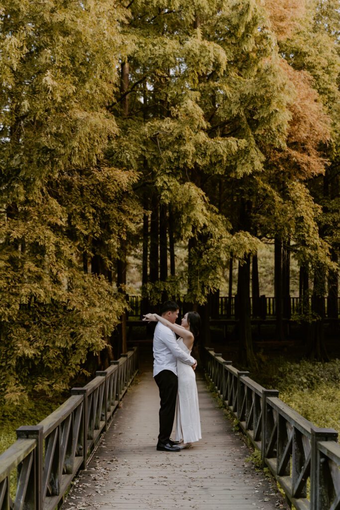 A couple embraces on a bridge in the woods.
