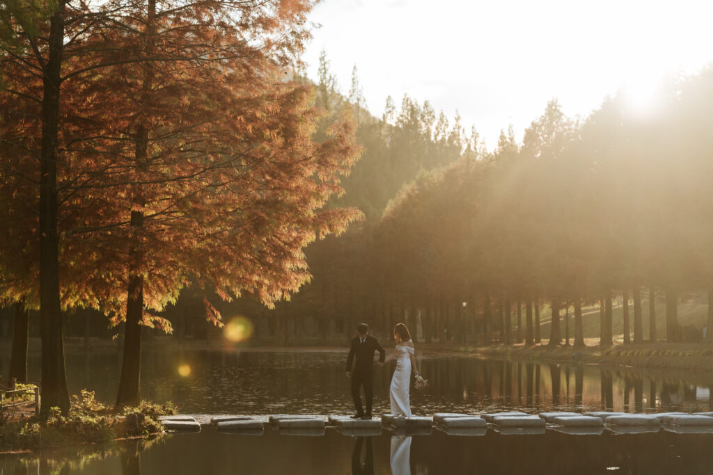 A bride and groom standing on a dock in front of a lake in damyang metasequoia road