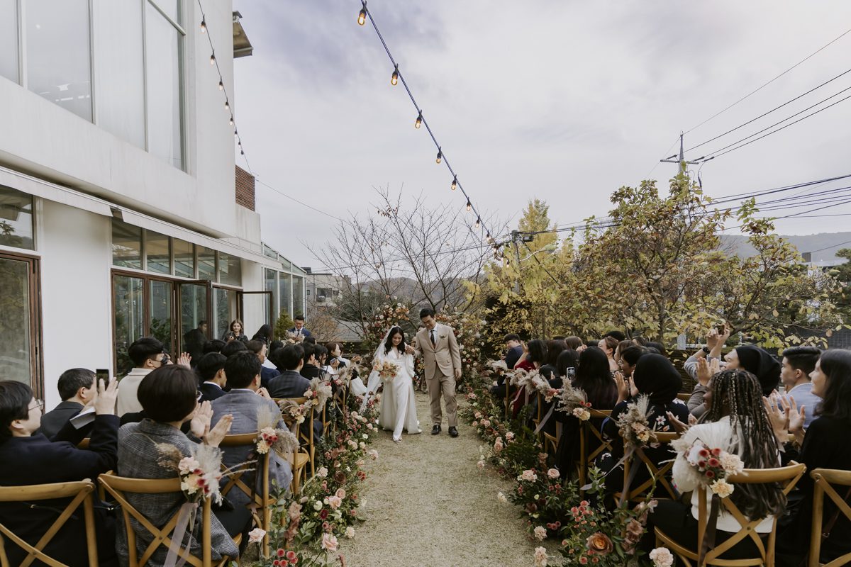 Bride and groom walking down the aisle at an outdoor wedding ceremony with guests seated on either side, surrounded by floral decorations and string lights.