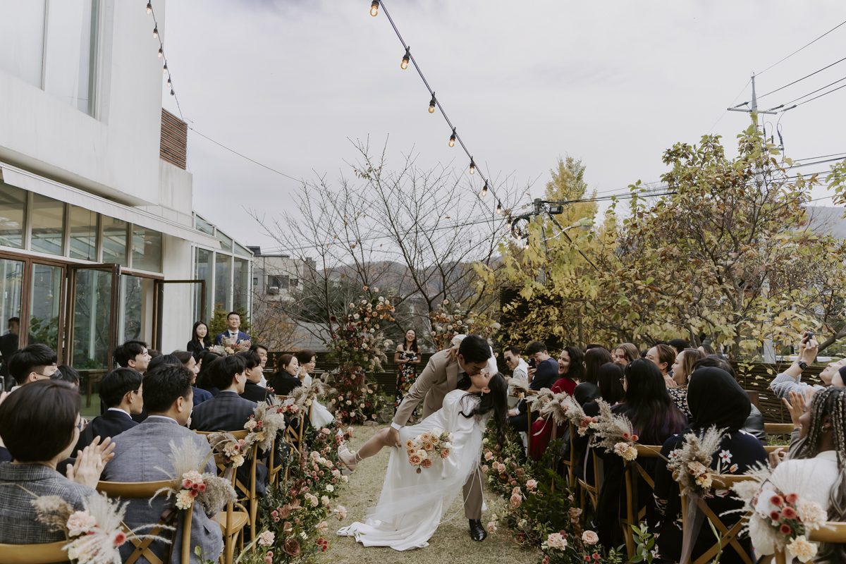 A couple kissing at the end of the aisle during an outdoor wedding ceremony, surrounded by seated guests and autumnal foliage.