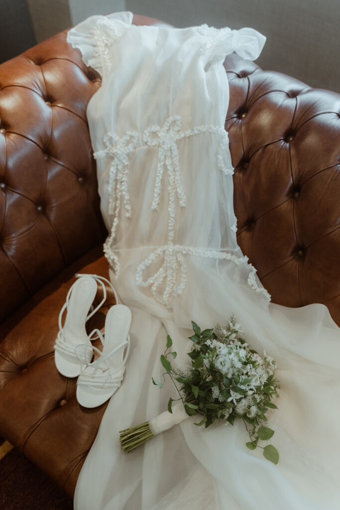 A white wedding dress with lace details lies on a brown tufted leather sofa, evoking the elegance of a Korean wedding. White sandals and a bouquet of white flowers with greenery are placed beside the dress.