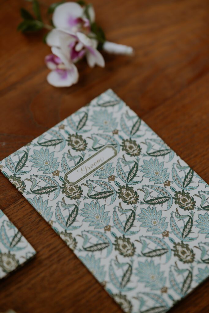 A floral-patterned booklet labeled "Groom" lays on a wooden surface with an orchid flower in the background.