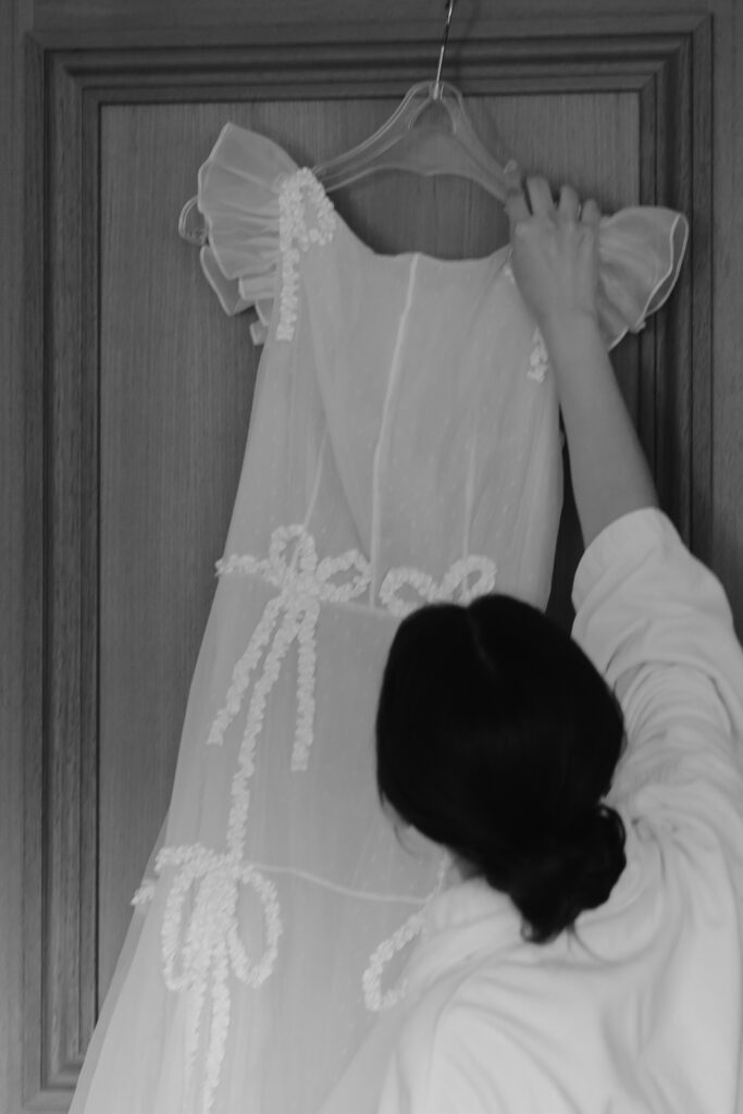A bride is reaching up to hang a white dress with delicate details on a wooden door.