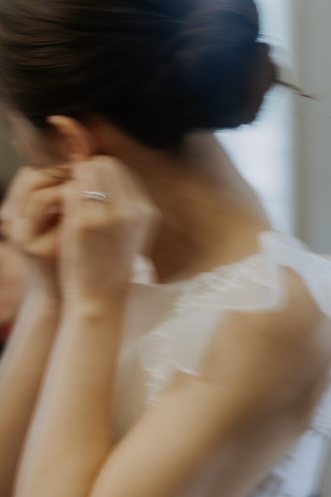 A woman with dark hair in a bun adjusts an earring. She is wearing a white, sleeveless dress.