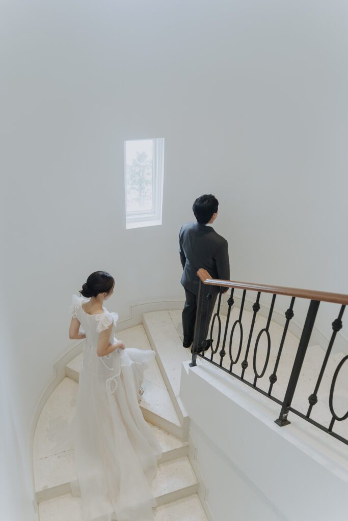 A bride in a white dress descends a curved staircase toward a groom in a dark suit standing at the base, both positioned in a minimalistic, light-filled space.
