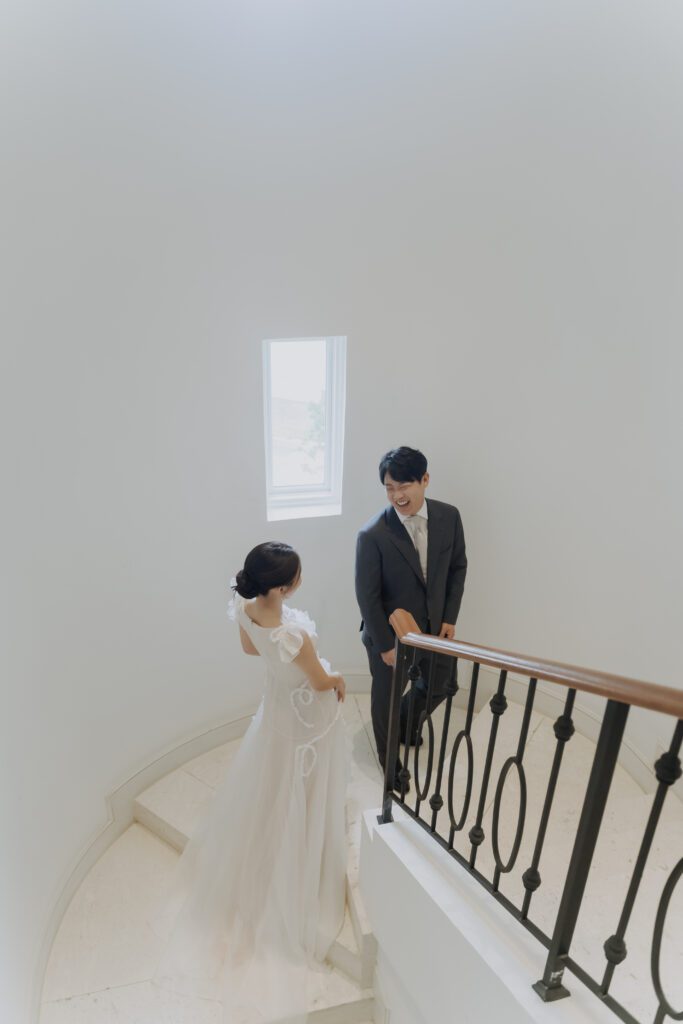 A couple dressed in formal attire stand on a curved staircase, facing each other. The woman is wearing a white gown, and the man is wearing a dark suit with a bow tie. A small window is above them.