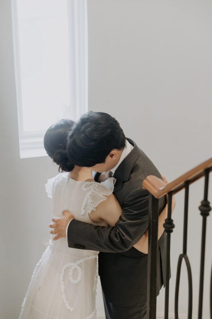A man in a suit and a woman in a white dress embrace on a staircase near a window with light streaming through, reminiscent of an enchanting scene from Korea.