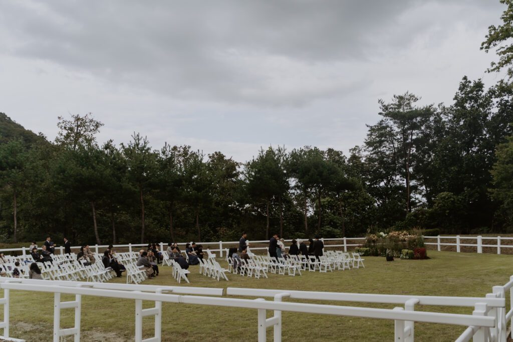 An outdoor Korean wedding ceremony with guests seated on white chairs in a grassy, fenced area surrounded by trees under a cloudy sky.