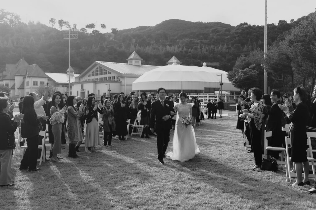 A bride and her father walk down an outdoor aisle lined with applauding guests during a beautiful Korean wedding ceremony, with a large building and trees in the background.
