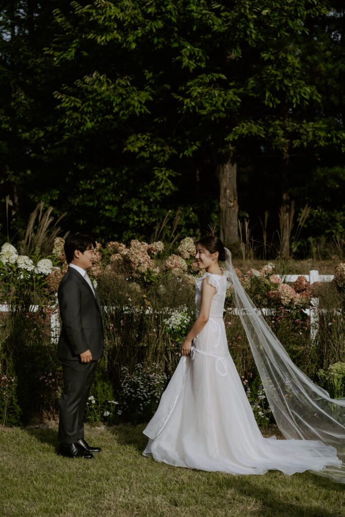 A couple stands facing each other in a garden, with the bride in a white gown and veil, and the groom in a dark suit. They are surrounded by greenery and flowers, creating an idyllic setting for their Korean wedding.