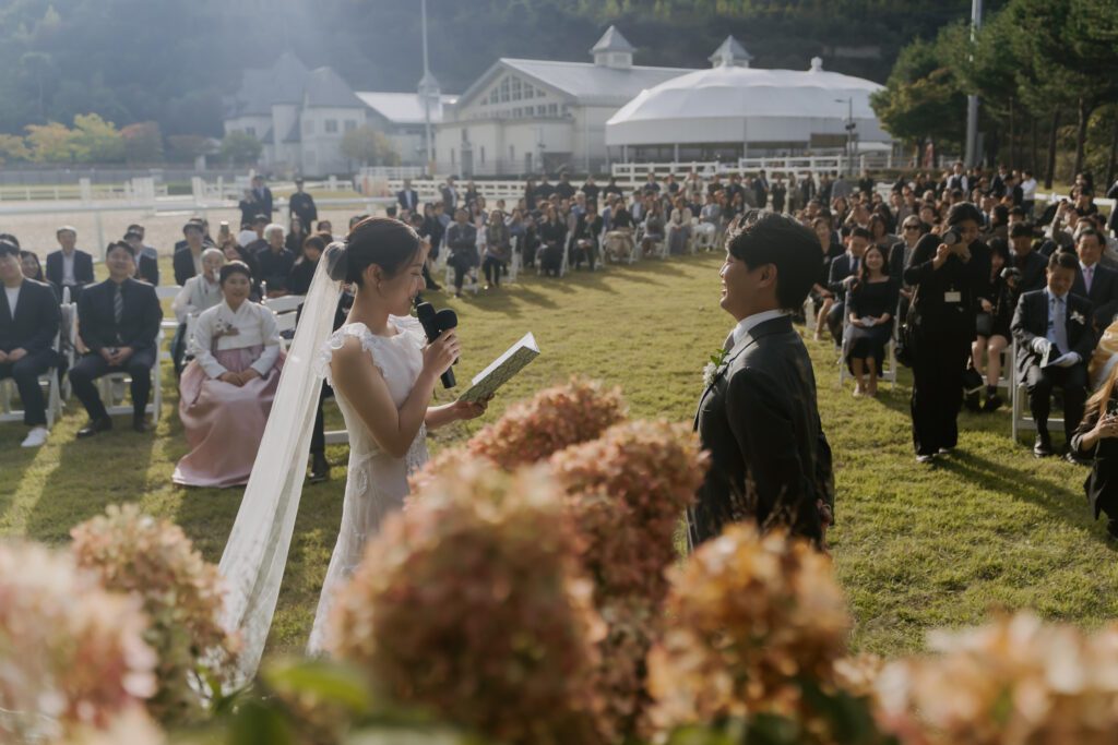 A bride and groom stand facing each other during their outdoor wedding ceremony, with guests seated in the background on a sunny day.