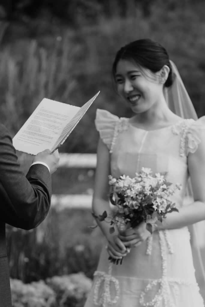 At a beautiful Korean wedding, a bride holding a bouquet of flowers smiles while listening to a person holding a piece of paper during the outdoor ceremony.