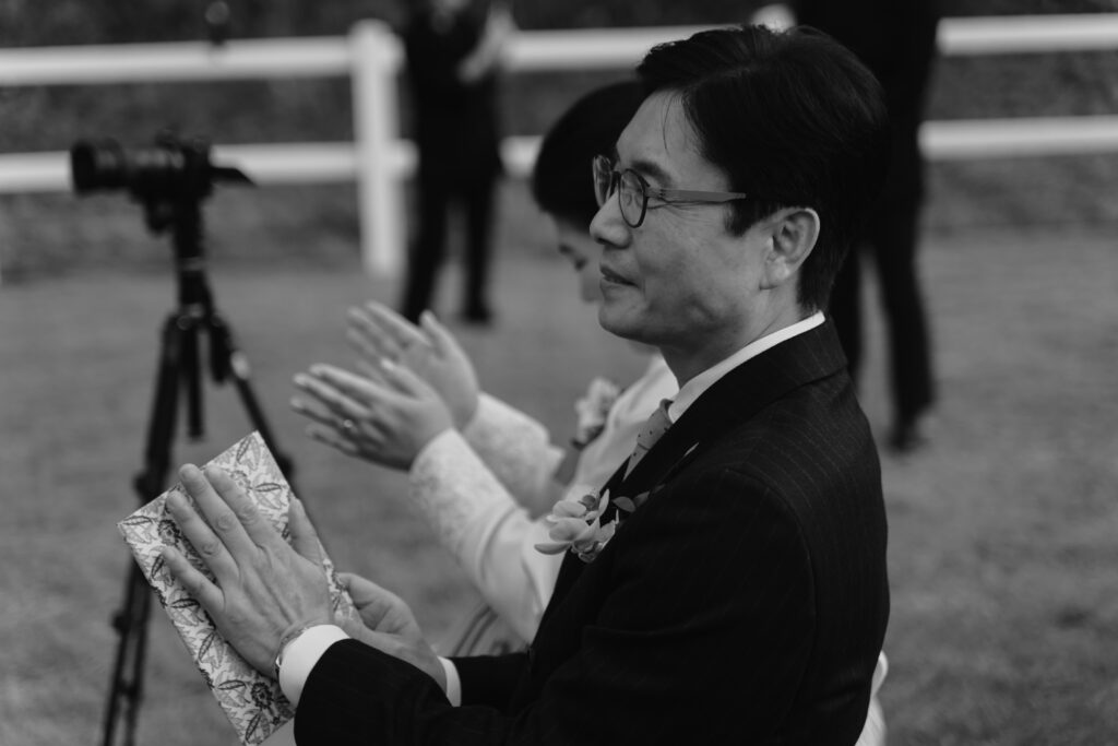 Black and white photo of two people clapping, seated outdoors. A man in the foreground holds a patterned envelope, reminiscent of a Korean wedding favor. A camera on a tripod is in the background.