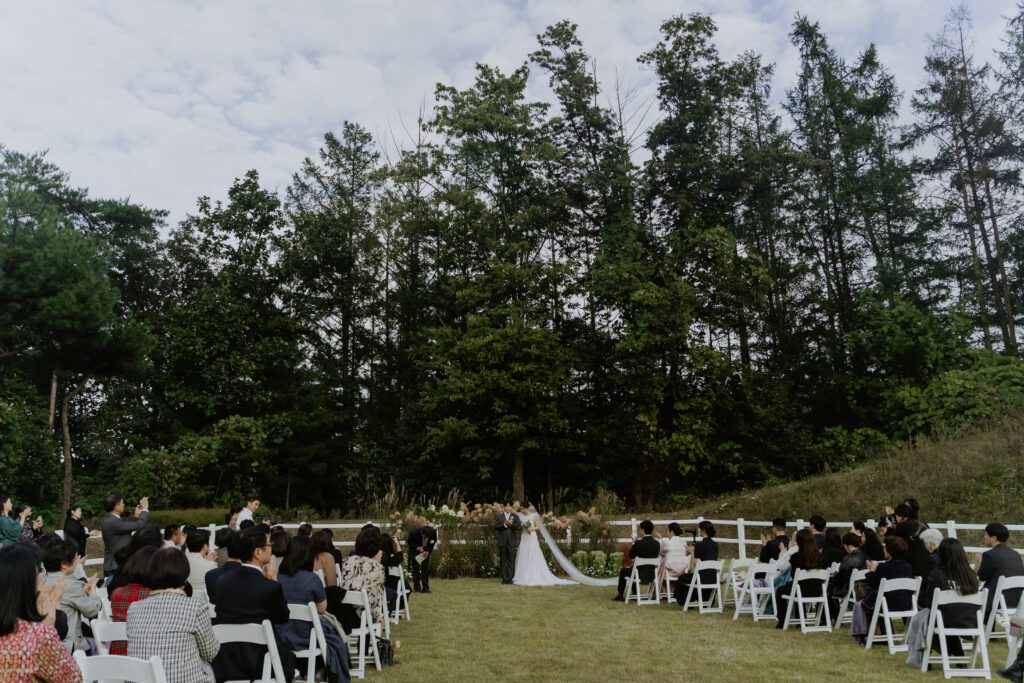 A Korean wedding ceremony taking place outdoors, with guests seated in white chairs facing a couple standing under an arch, surrounded by trees and greenery.