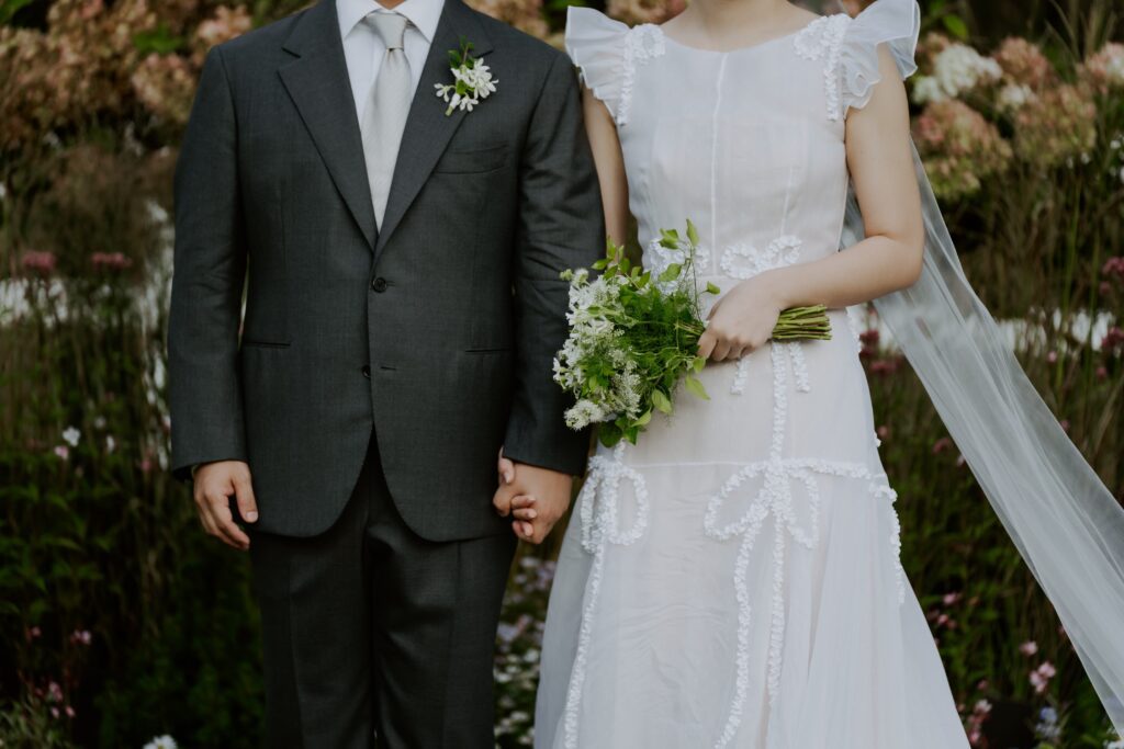 A bride and groom holding hands at their Korean wedding; the groom is wearing a dark suit with a boutonnière, while the bride dons a white dress with a bouquet in hand, her veil trailing behind her. The background features vibrant foliage.