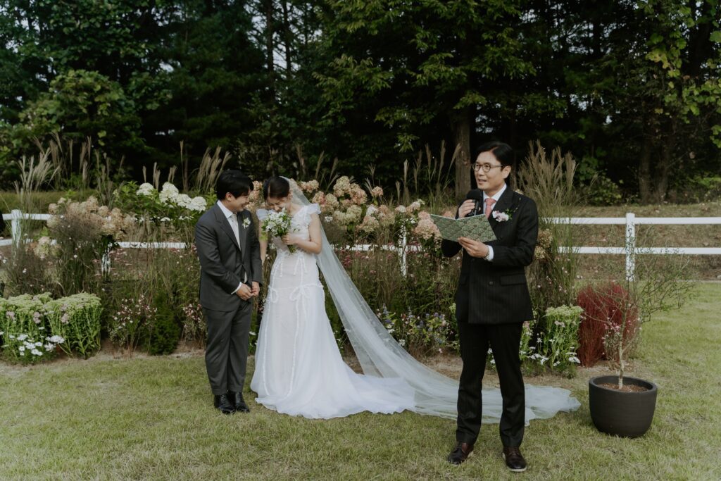 A couple, dressed in wedding attire, stands close together outdoors beside the father of the bride who is reading from a book. The scene is reminiscent of a Korean wedding, set against lush greenery and a pristine white fence.