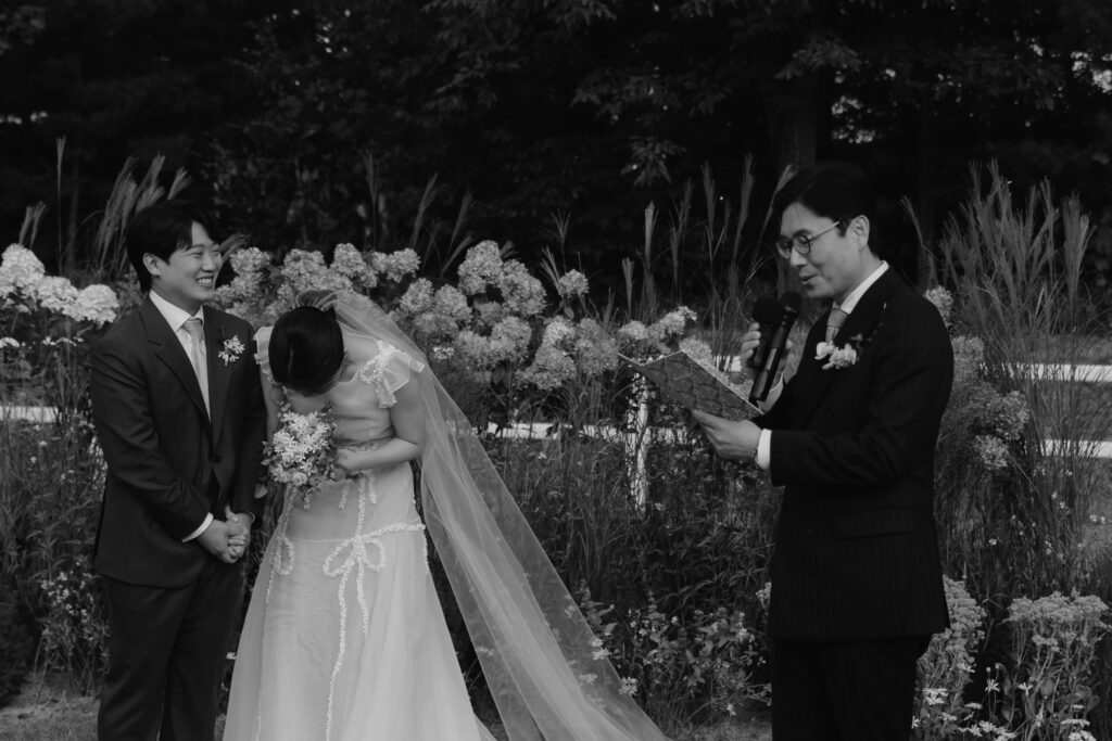 A couple stands in front of a person reading from a book, surrounded by flowers at their Korean wedding. The woman is laughing, holding a bouquet, while the man is smiling.