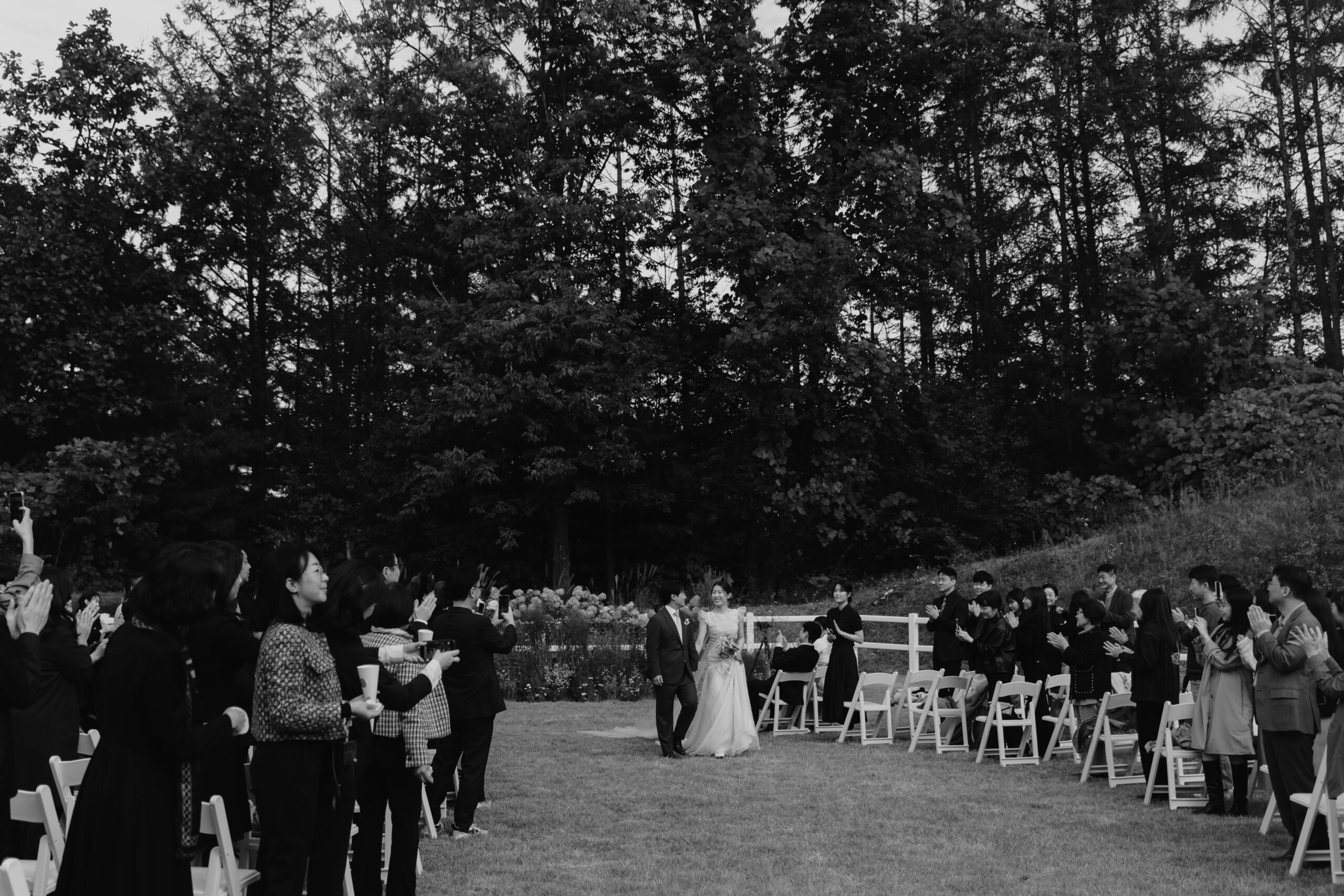 A couple in wedding attire walks down the aisle outdoors, surrounded by seated guests who are clapping and standing in an aisle lined by chairs. Trees and greenery serve as the backdrop to this enchanting Korean wedding.