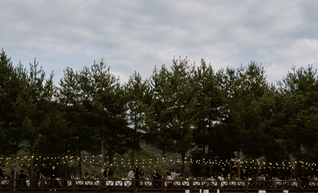 Outdoor reception on wooden deck with string lights, set against a backdrop of tall evergreen trees and a cloudy sky, reminiscent of an intimate Korean wedding.