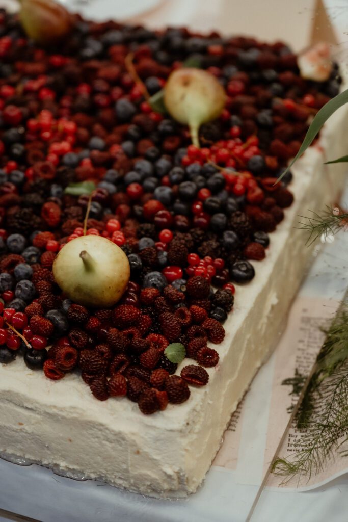 A rectangular cake topped with a variety of berries and whole figs, perfect for a Korean wedding. The cake is adorned with white frosting and decorated with sprigs of greenery on the sides.