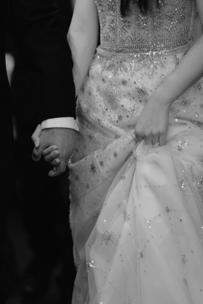 A couple holding hands at a Korean wedding; the woman wears an embellished gown and holds up its fabric while the man wears a suit.
