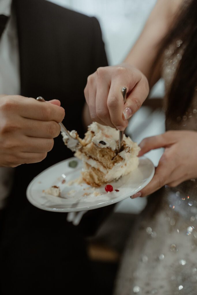 Two people sharing a slice of cake on a white plate, each holding a fork. Dressed formally, the scene looks like it could be from an elegant Korean wedding.