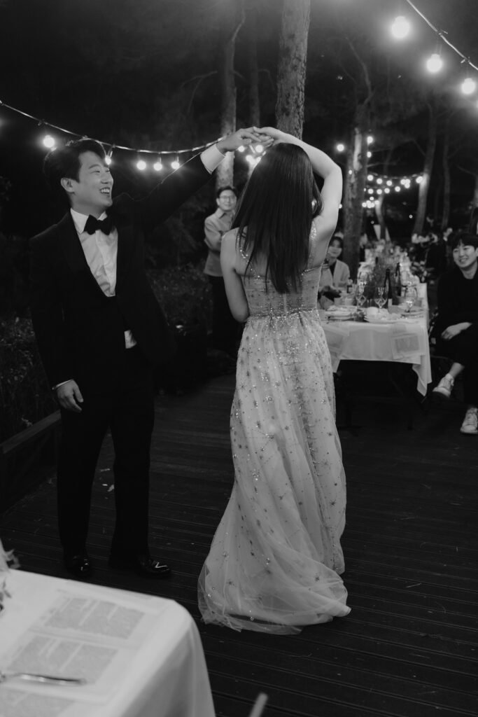 A couple dances outdoors under string lights at a nighttime Korean wedding. The man wears a suit while the woman wears a long dress. Guests sit at tables in the background.