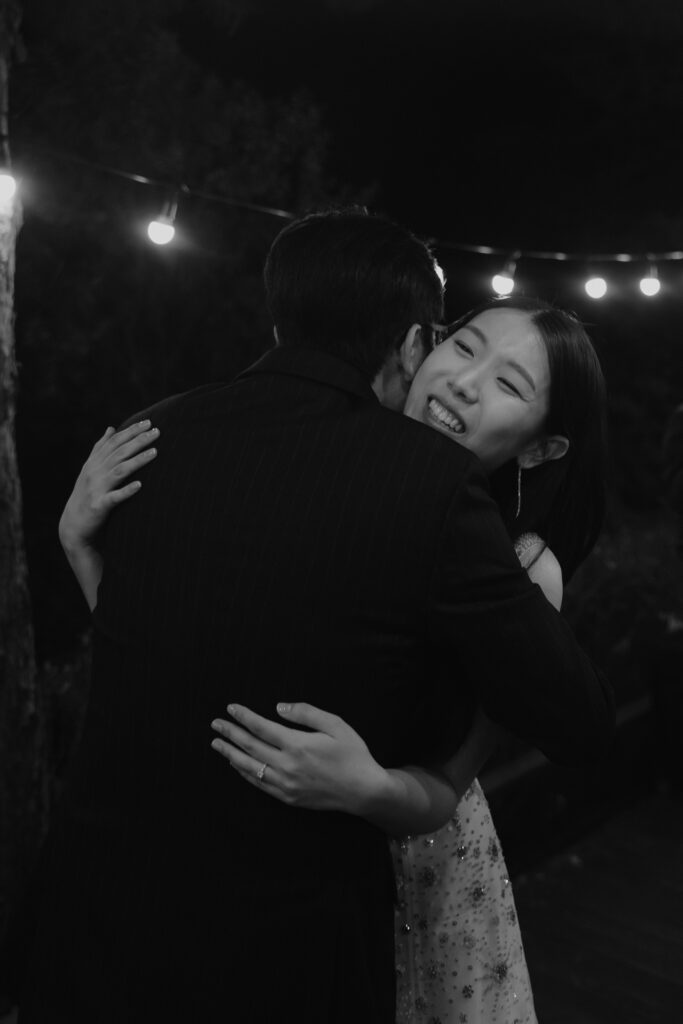 Two people share a hug under string lights at night. The person facing the camera is smiling while wearing a light-colored, sleeveless dress. The other person, dressed in dark attire with their back to the camera, adds to the intimate moment reminiscent of a romantic Korean wedding scene.