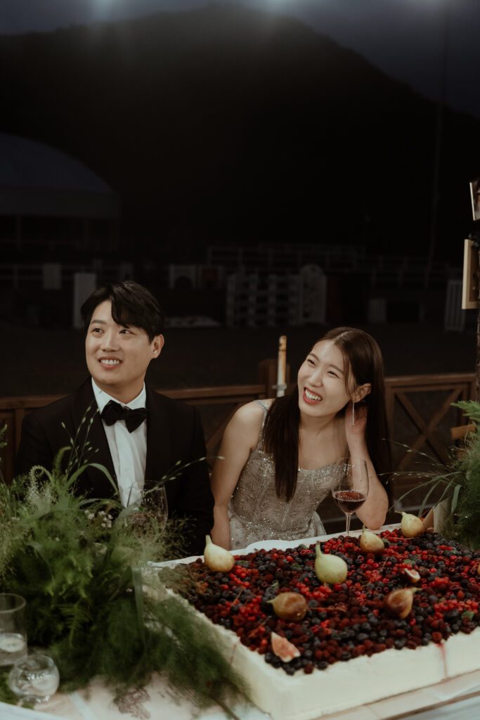 A man and woman sitting at a table with a large cake covered in berries and figs in front of them, reminiscent of a traditional Korean wedding. The woman is smiling and holding a glass, while the man is looking to the side.