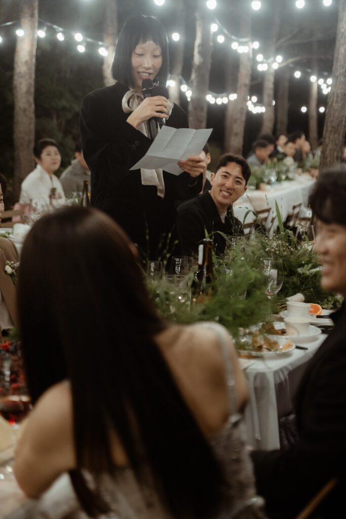 A woman stands and reads from a paper while speaking into a microphone at a formal Korean wedding dinner event with string lights and trees in the background. Guests at the table are listening attentively.