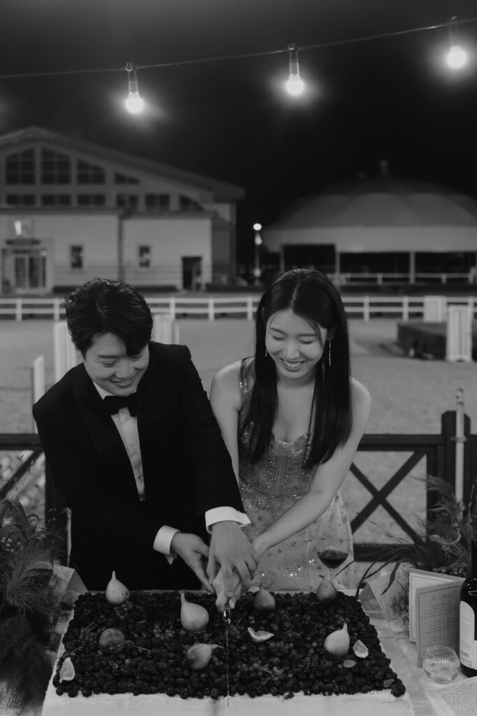 A man in a suit and a woman in a dress smile as they cut into a large cake covered in fruit. They are outside at night under string lights, celebrating their lovely Korean wedding.