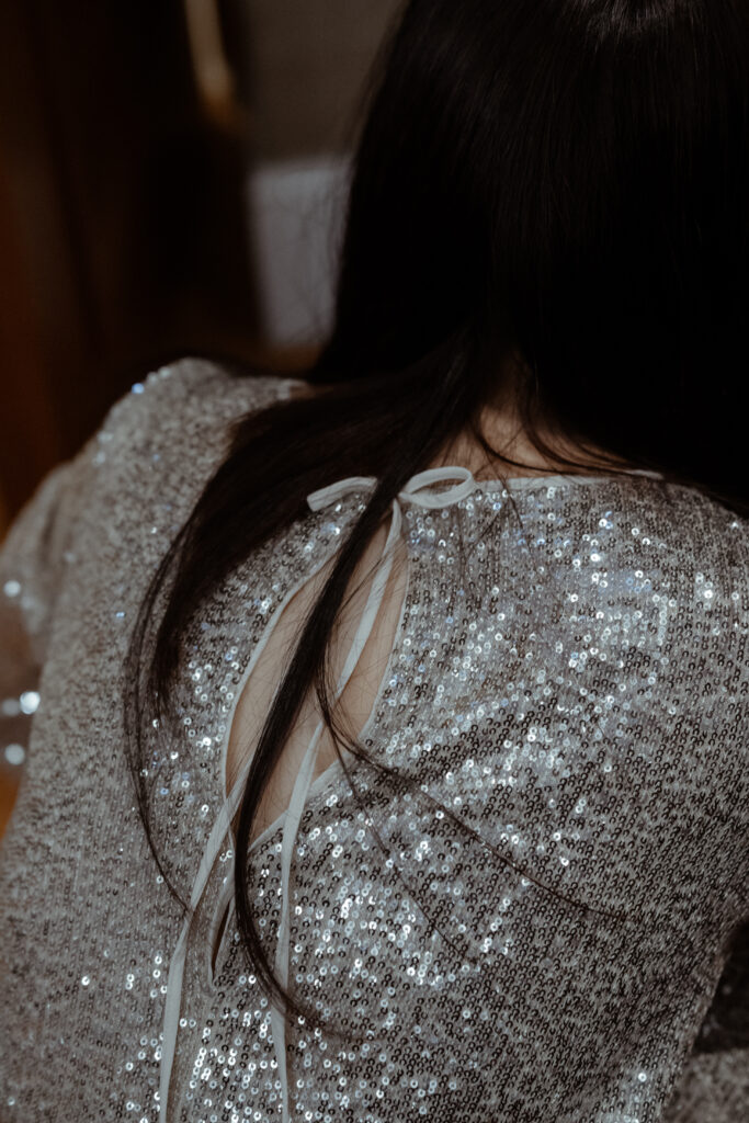 A person with long dark hair is shown from behind, wearing a glittery silver top with an opening tied with a ribbon at the back, evoking the elegance of a Korean wedding.