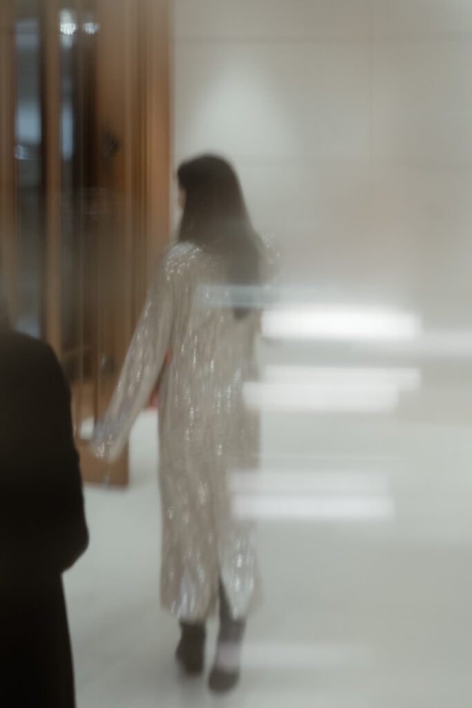 A person with long dark hair is walking away, wearing a long, shiny coat reminiscent of traditional Korean wedding attire. The image is blurry with light streaks and reflections.